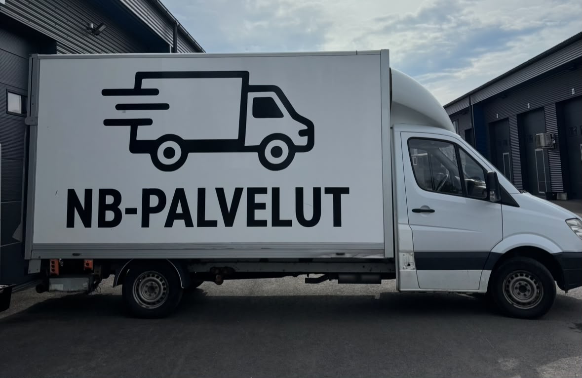 Valkoinen kuorma-auto, jossa kyljessä NB-PALVELUT-teksti ja lavakuva, varastohallien edessä.
