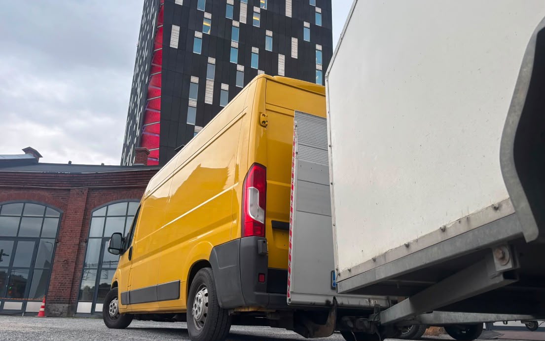 Keltainen pakettiauto lastauslavalla, perävaunun kylki näkyy, taustalla korkea toimistorakennus ja tiiliseinä.